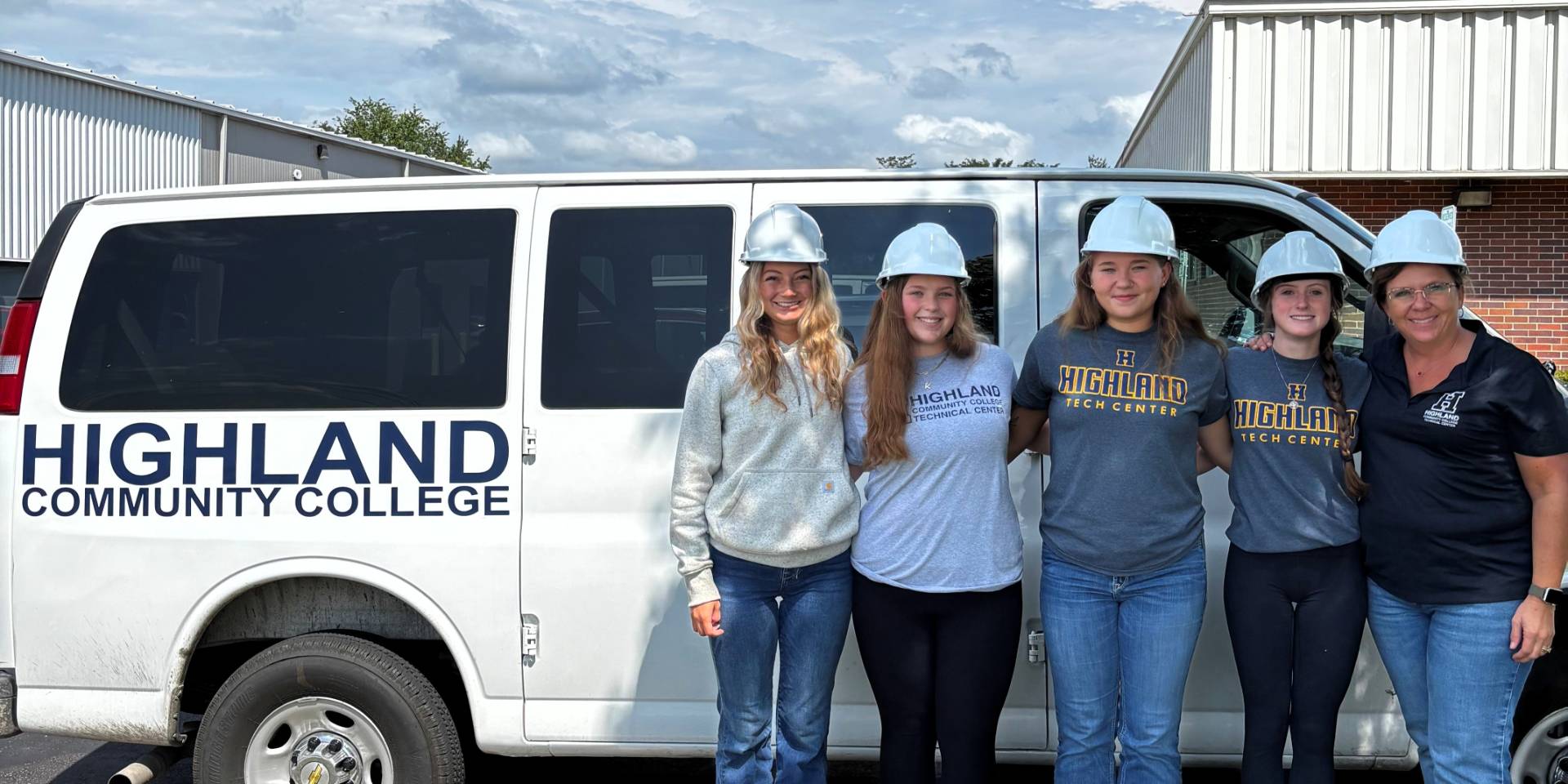 Group of students posing in front of a van with a decal "Highland Community College." Students are wearing hard hats.