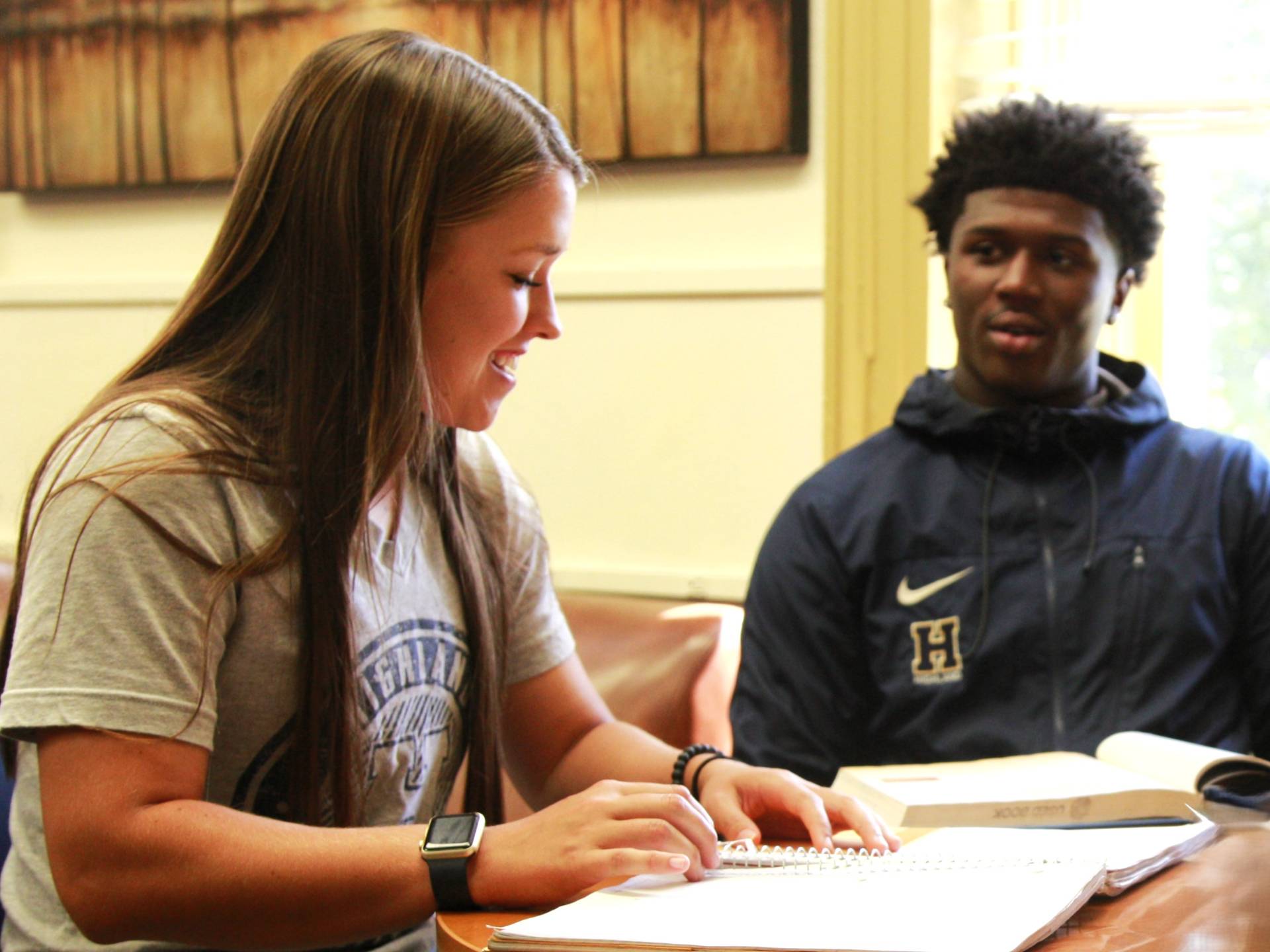 Two students studying at a table, one student is looking at a notebook