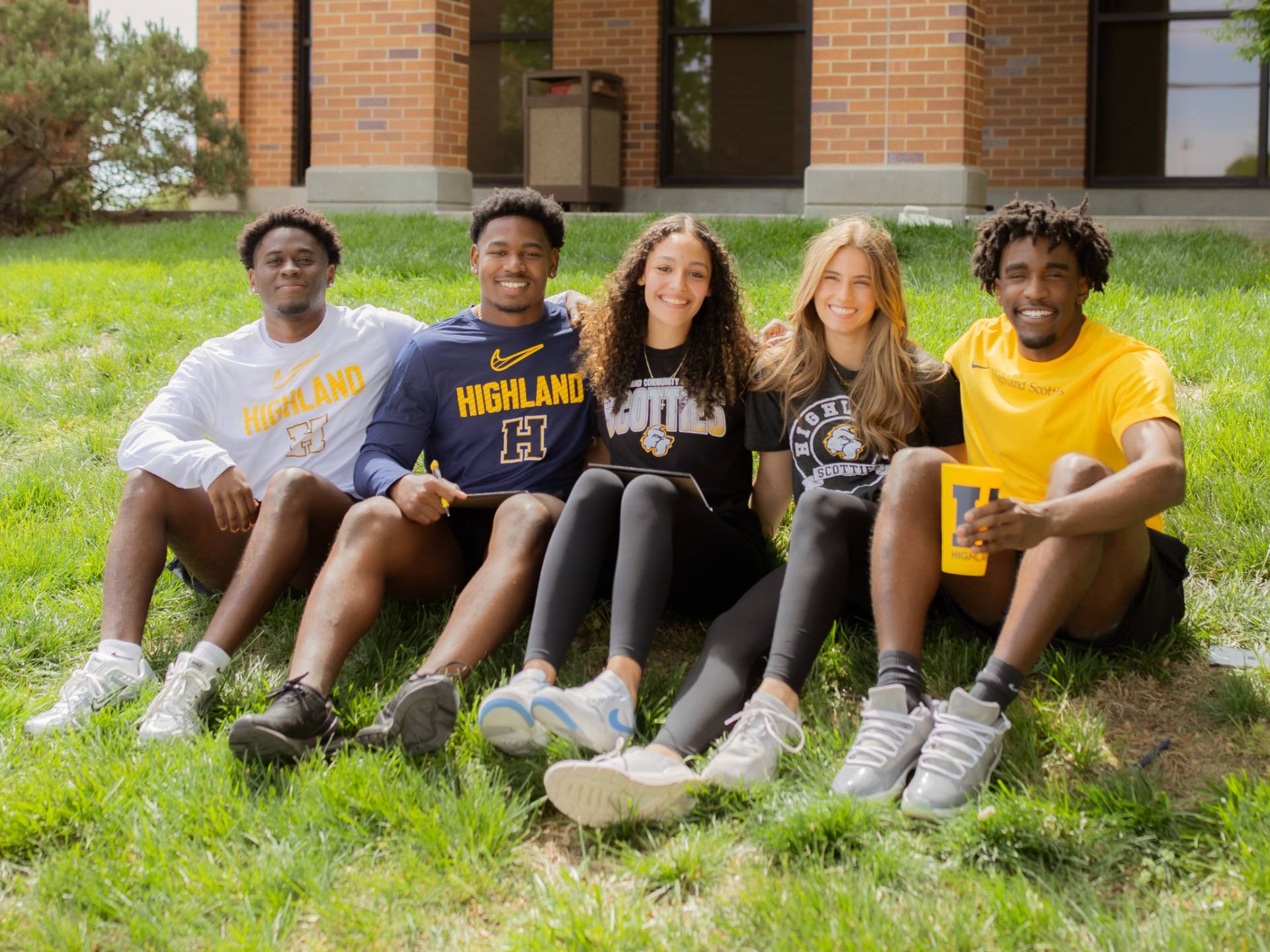 Five students sitting on the lawn in a line smiling at the camera.