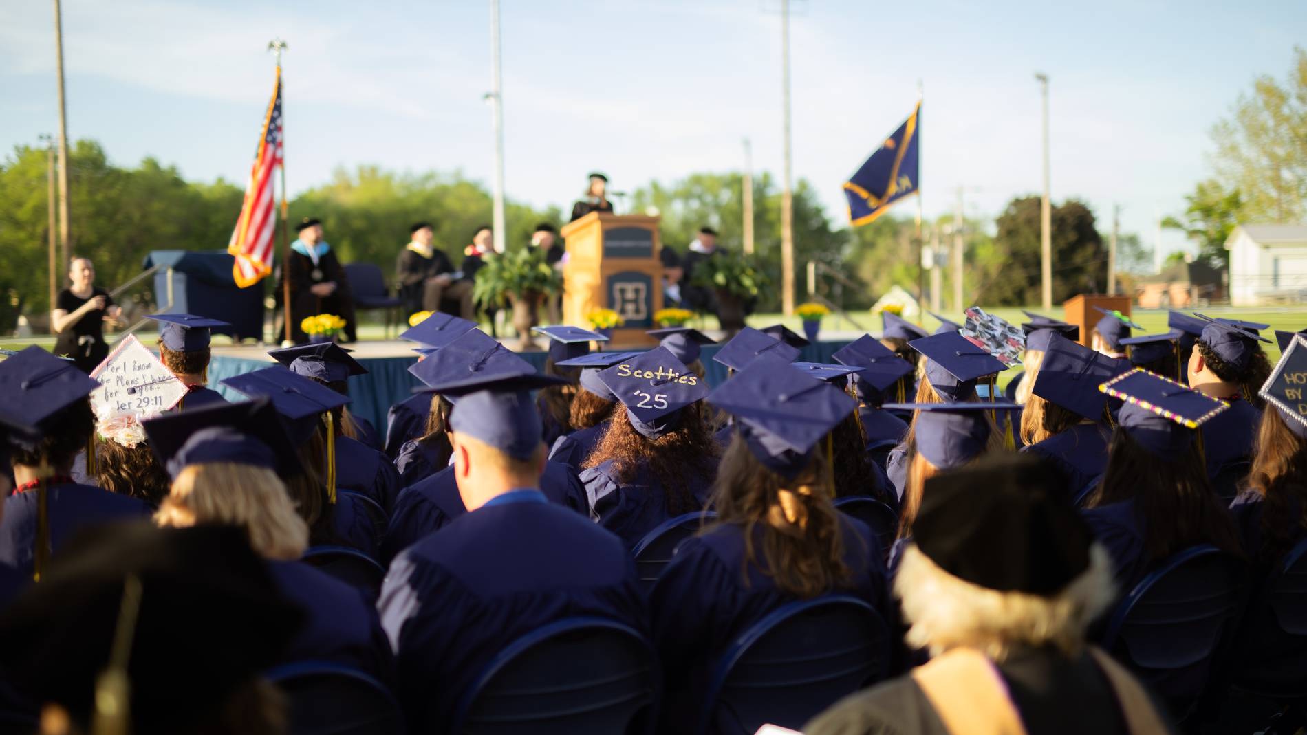 Graduates sit in chairs on the football field, facing the podium speaker.